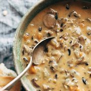 White hand holding a spoon in a bowl of Wild Rice Soup. There's broken crusty bread by the soup bowl.